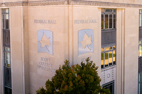 Aerial image of the corner of a large square building with a large metallic indiana state university sycamore leaf logo and the words “federal hall” above the leaf and “scott college of business” below the leaf with green trees in the foreground and on the side of the building.