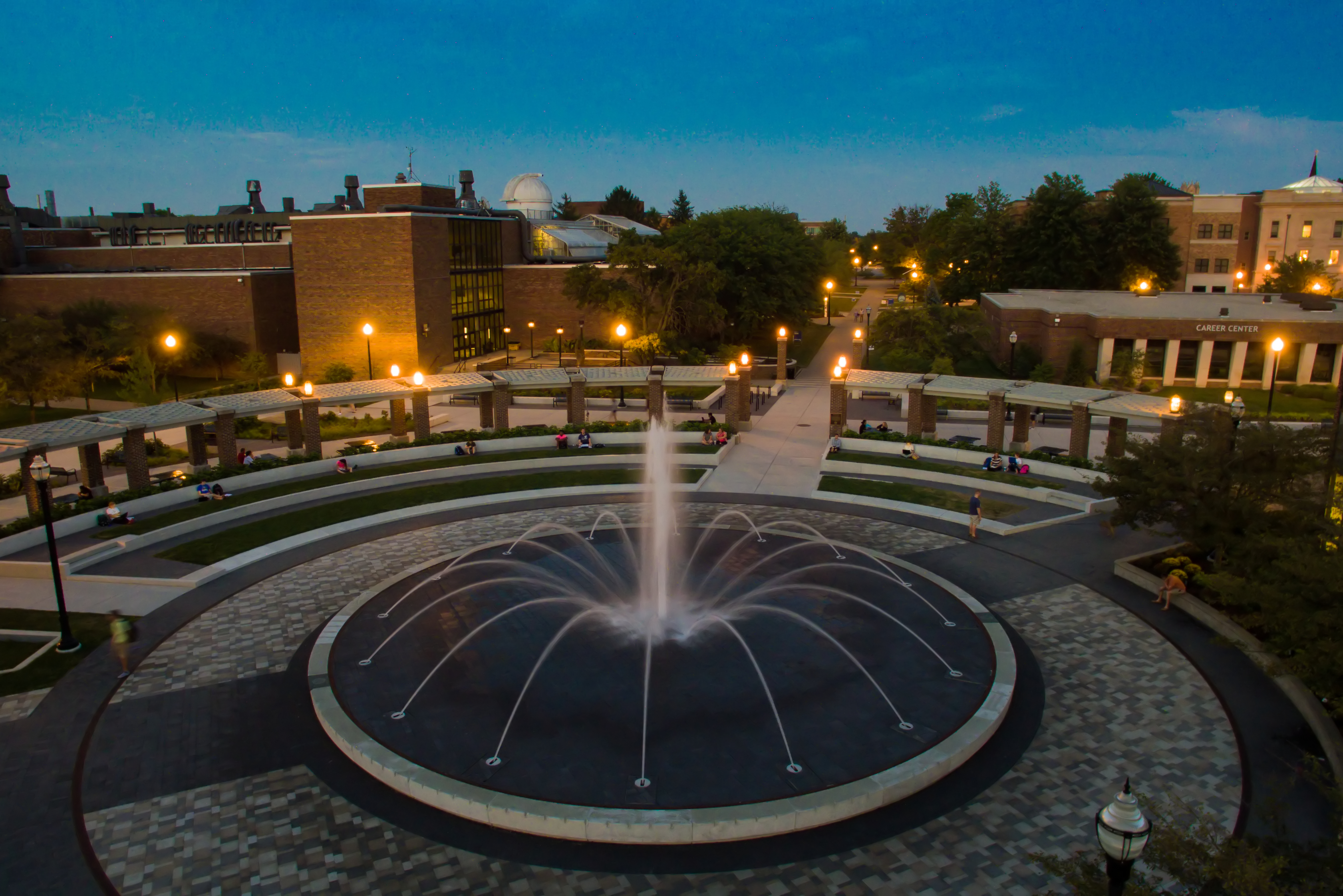 Aerial view of a circular fountain with illuminated jets of water at dusk, surrounded by brick buildings, walkways, and campus greenery.
