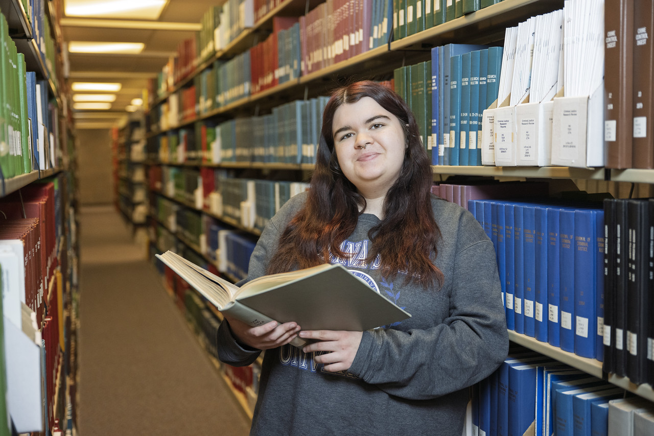 A white female with long brown hair, wearing a gray sweatshirt, is standing in a library aisle filled with bookshelves on both sides. She is holding an open book. The bookshelves are neatly organized with various colored books.
