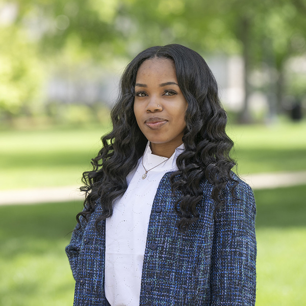 Shanisa Stinson stands smiling outdoors in a campus setting. She has long, curly black hair and is wearing a white shirt and a blue textured jacket. The background features green grass, trees, and blurred foliage.