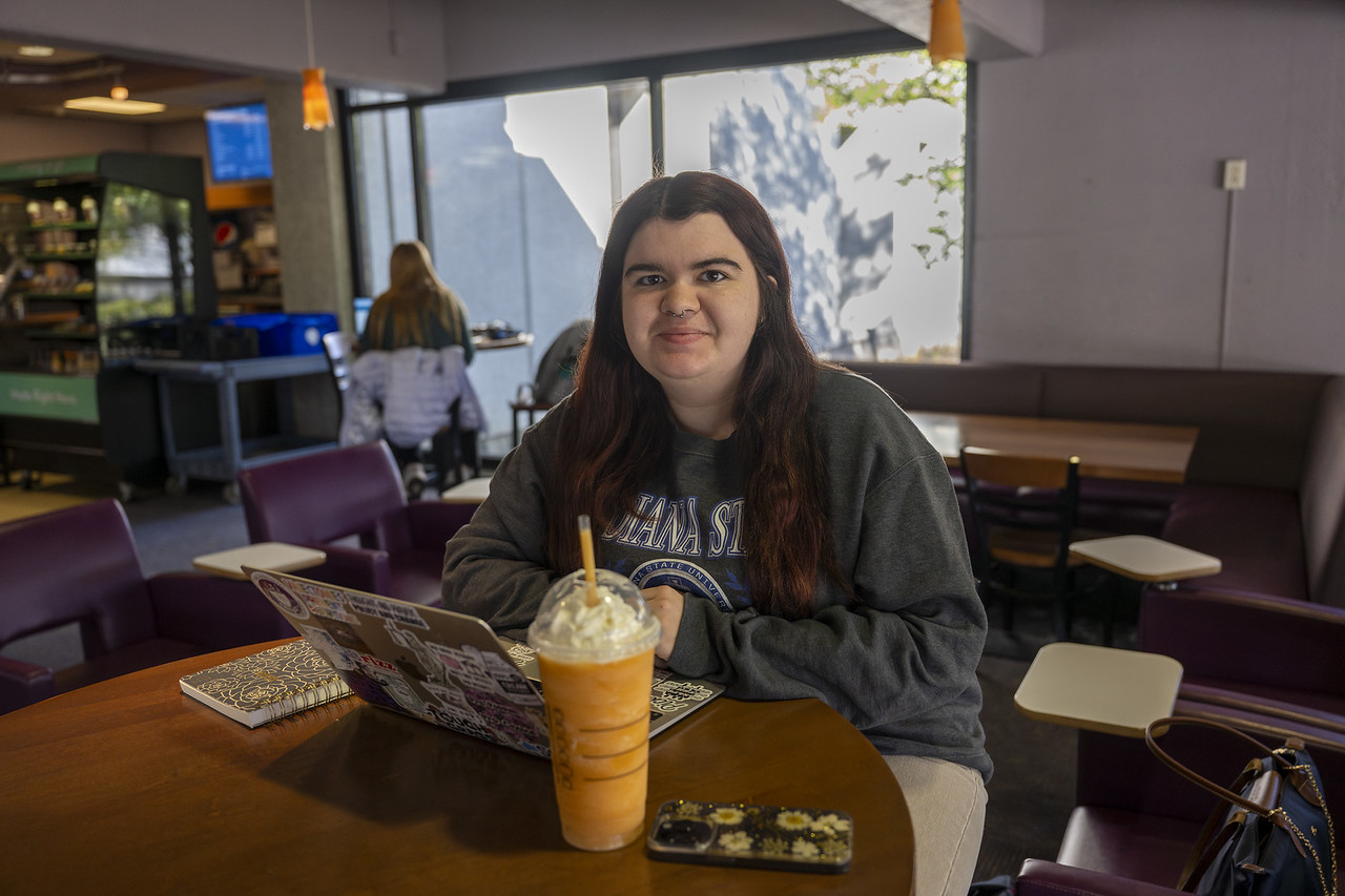 A young white woman with long dark hair sits at a table in a café, wearing a gray Indiana State University sweatshirt. In front of her are a laptop, a notebook, a phone, and a large orange Starbucks drink topped with whipped cream.