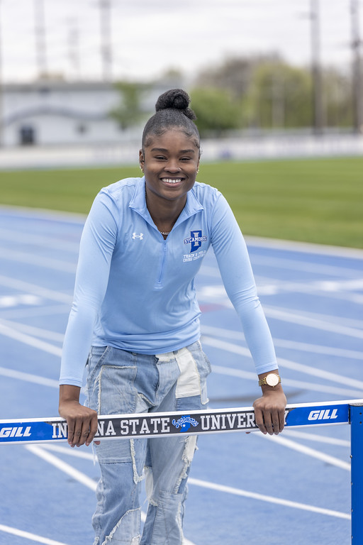 A young Black woman in a light blue long-sleeve shirt with an Indiana State Sycamores logo and patchwork jeans smiles while leaning on a hurdle that reads “Indiana State University.” A blue running track and grassy field are in the background.