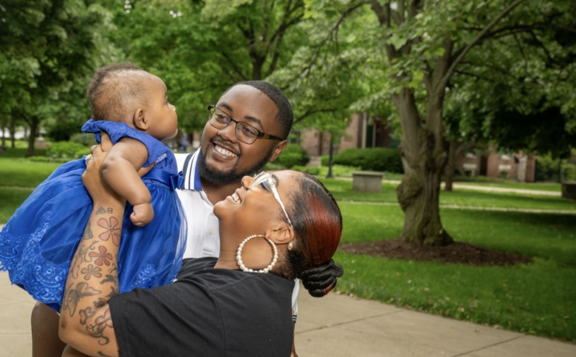 A smiling Black couple stands outdoors holding their baby dressed in a bright blue dress. The woman, wearing a black T-shirt, large hoop earrings, and clear glasses, lifts the baby toward her face, while the man, in a white collared shirt with blue trim and black glasses, looks on with a joyful expression. Green trees and walkways appear in the background.