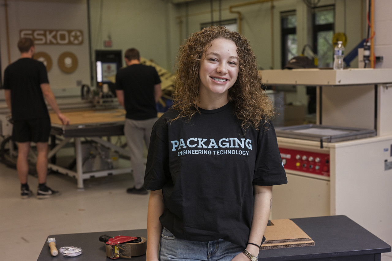 A female with curly brown hair is smiling and standing in a workshop, wearing a black t-shirt that reads 'PACKAGING ENGINEERING TECHNOLOGY.' In the background, three other individuals are engaged with machines and equipment. The workshop contains tools, machinery, and workbenches, creating an industrious, technical atmosphere.