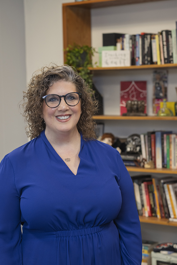A white woman with curly light brown hair and glasses, wearing a royal blue blouse, stands in front of a bookshelf filled with books, decorative items, and a small green plant.