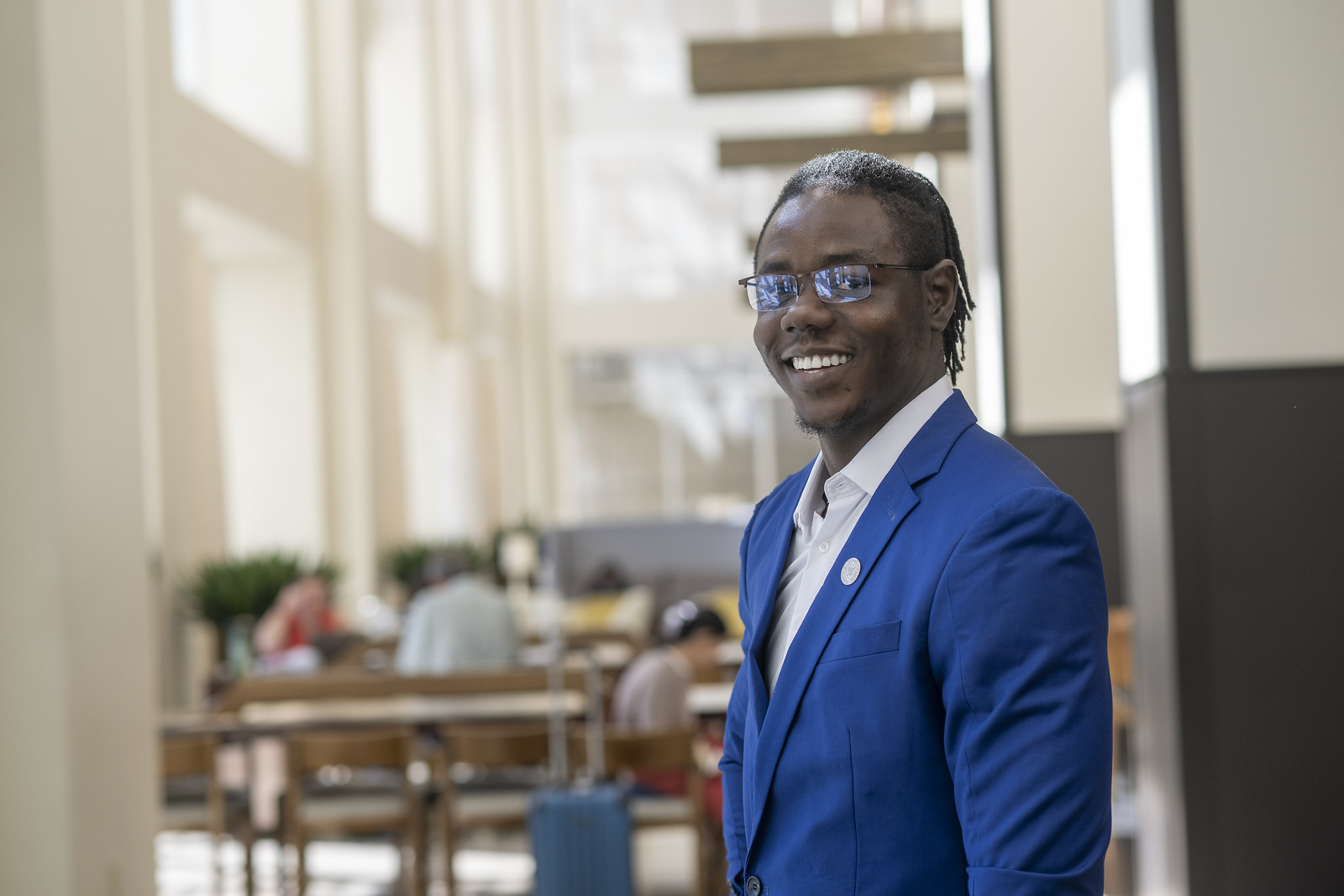 A Black man with braided hair, wearing a bright blue suit over a white shirt and glasses, stands in a modern, sunlit building with people seated in the background.