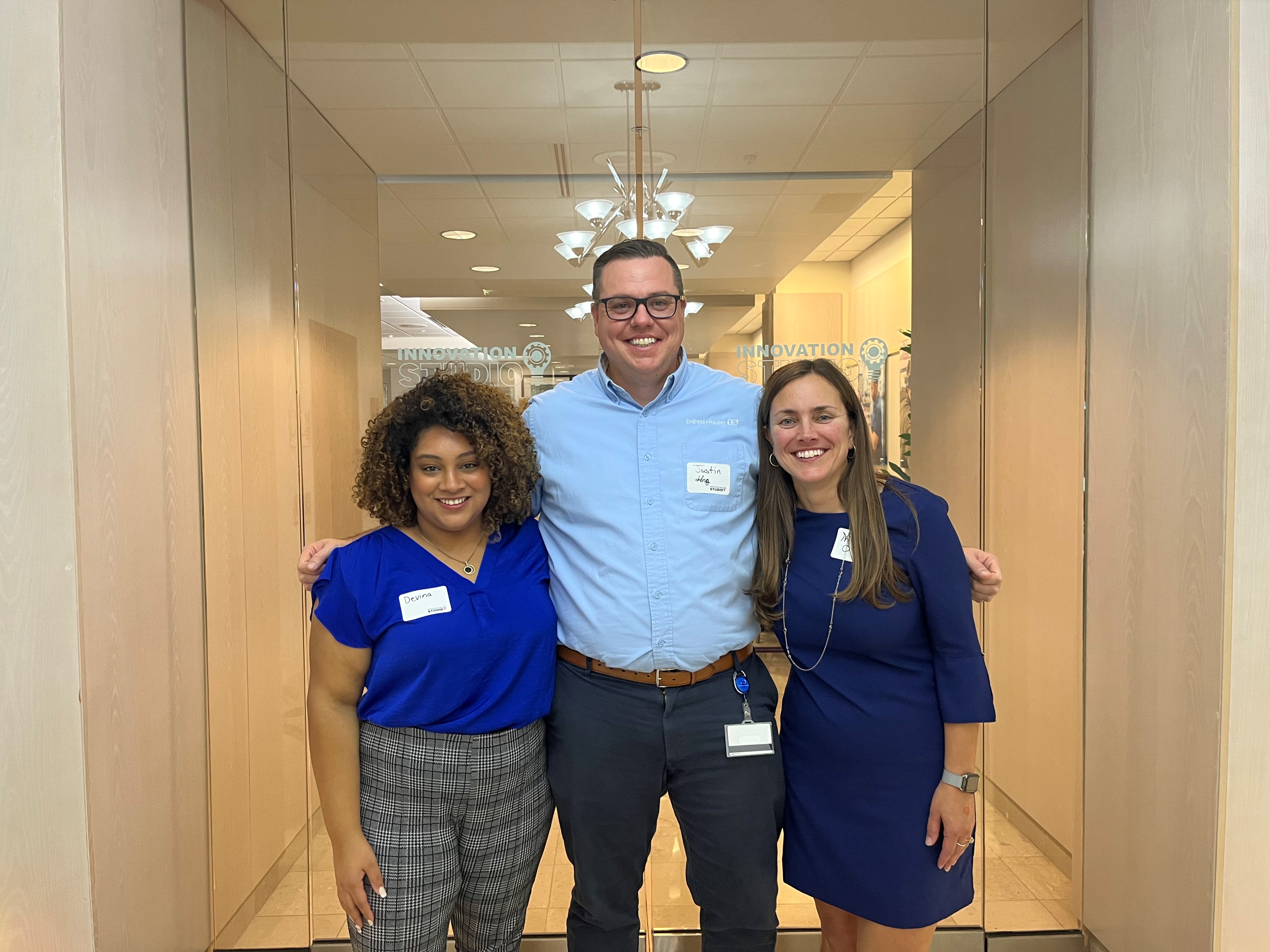 A Black woman in a blue top and plaid pants, a White man in a light blue shirt and dark pants, and a White woman in a blue dress stand smiling in a bright hallway lined with glass walls. Each wears a name tag, and signs reading “INNOVATION” and “INTERNATIONAL” are visible behind them.