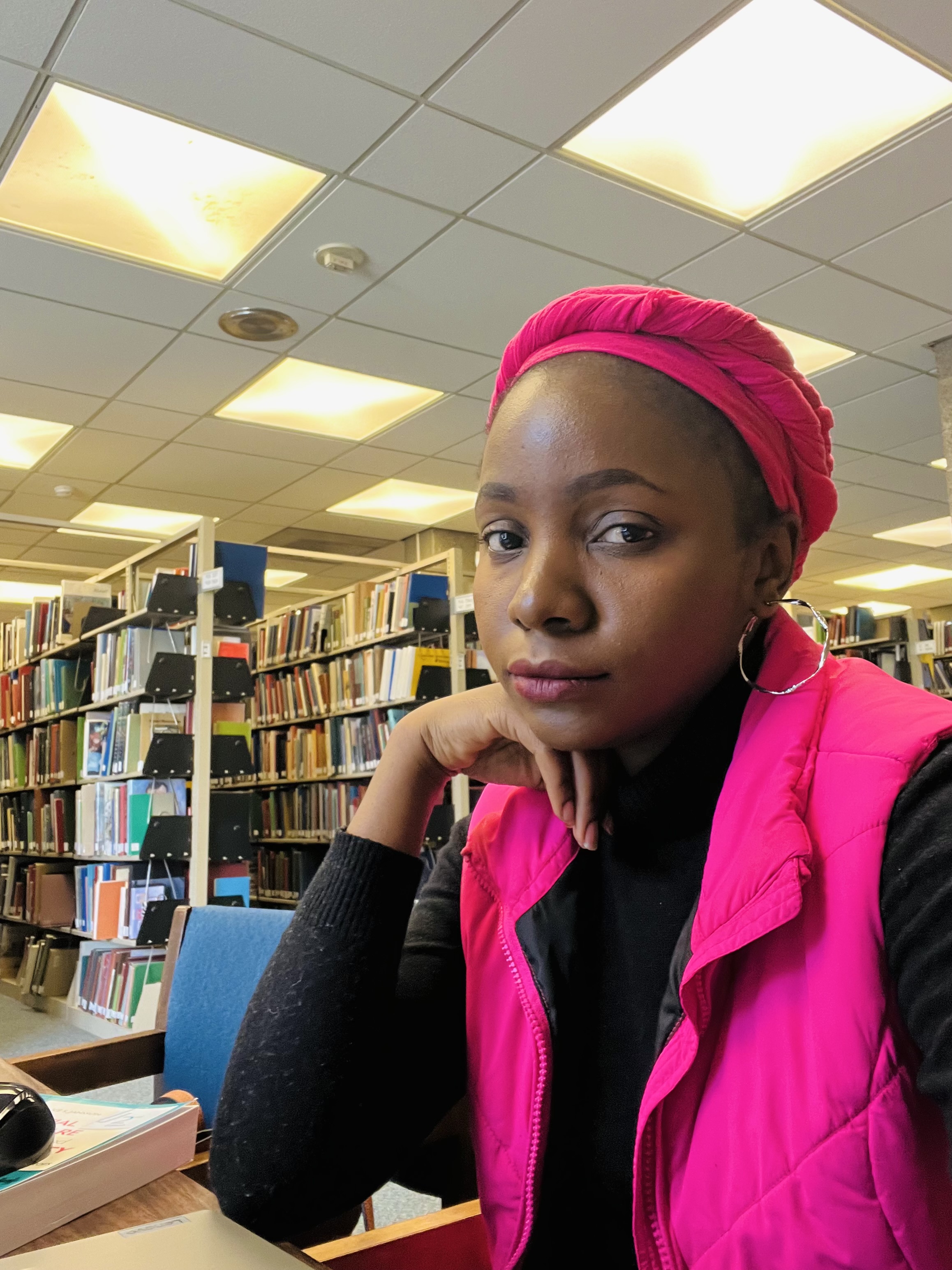 A young Black woman wearing a bright pink headwrap and matching vest sits in a library, resting her chin on her hand. She wears a black long-sleeve top and hoop earrings, with bookshelves and ceiling lights in the background.