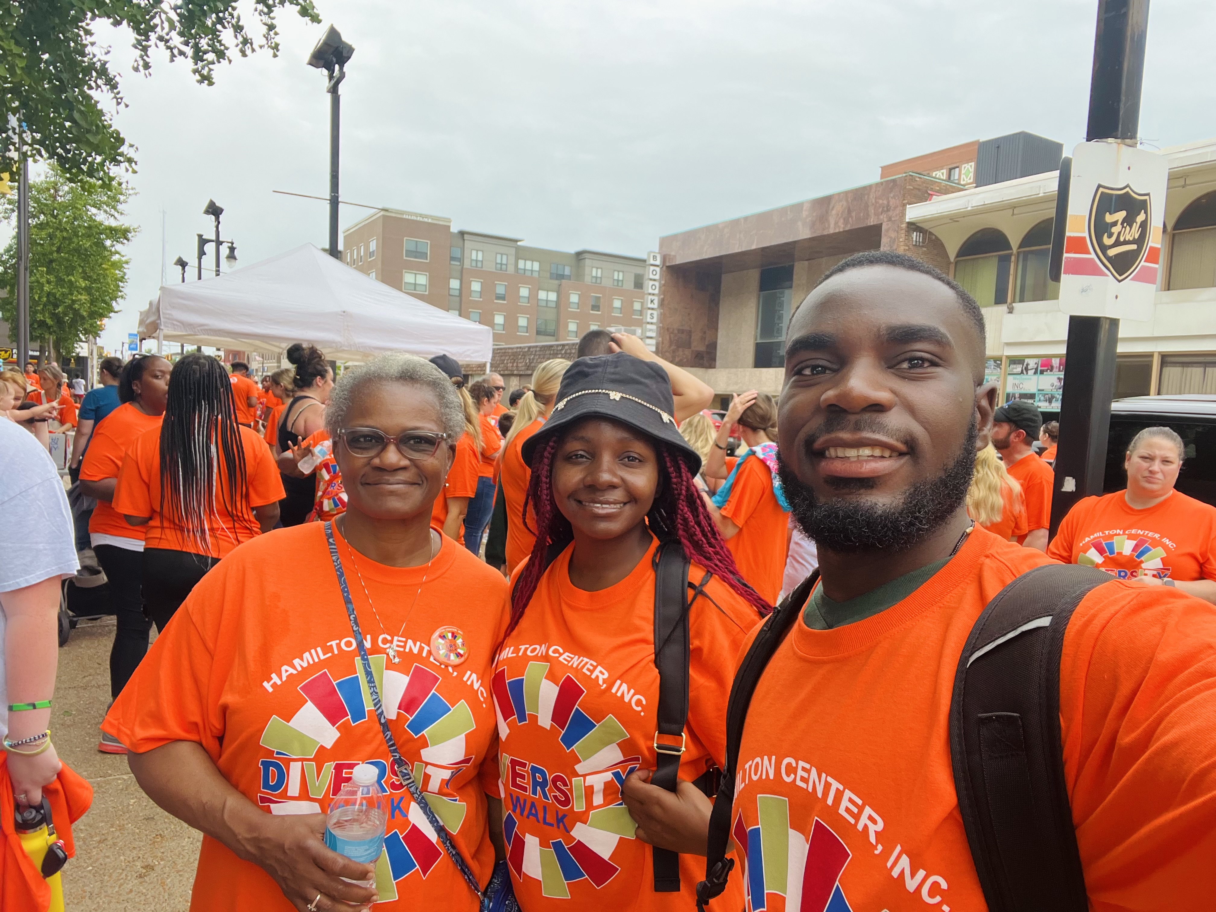 Three smiling Black individuals stand together at an outdoor event wearing bright orange “Hamilton Center, Inc. Diversity Walk” T-shirts. The older woman on the left holds a water bottle, the young woman in the middle wears a black bucket hat, and the man on the right carries a backpack.