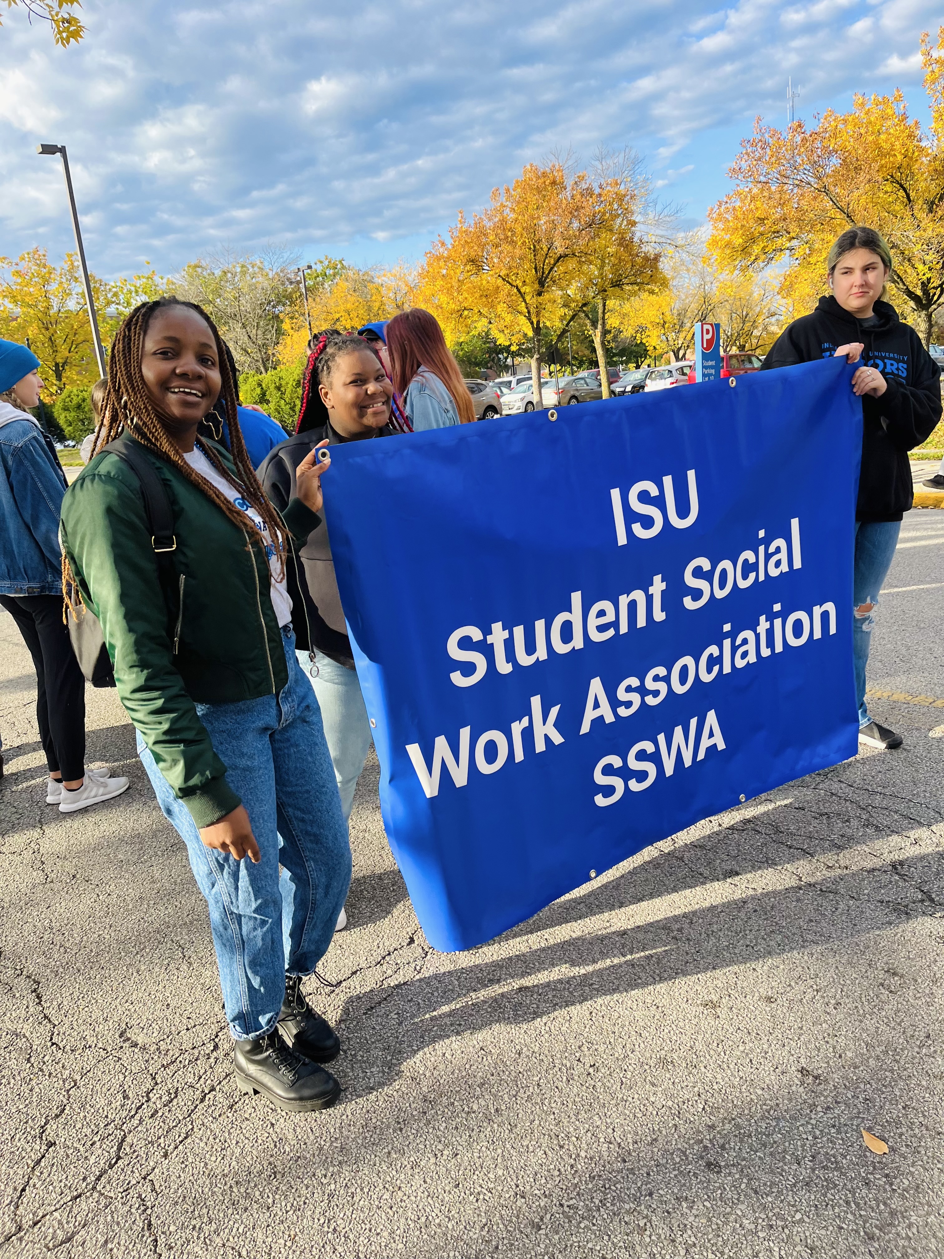 Three young women stand outdoors holding a blue banner that reads “ISU Student Social Work Association SSWA.” The woman on the left, who is Black, smiles while wearing a green jacket, jeans, and black boots.