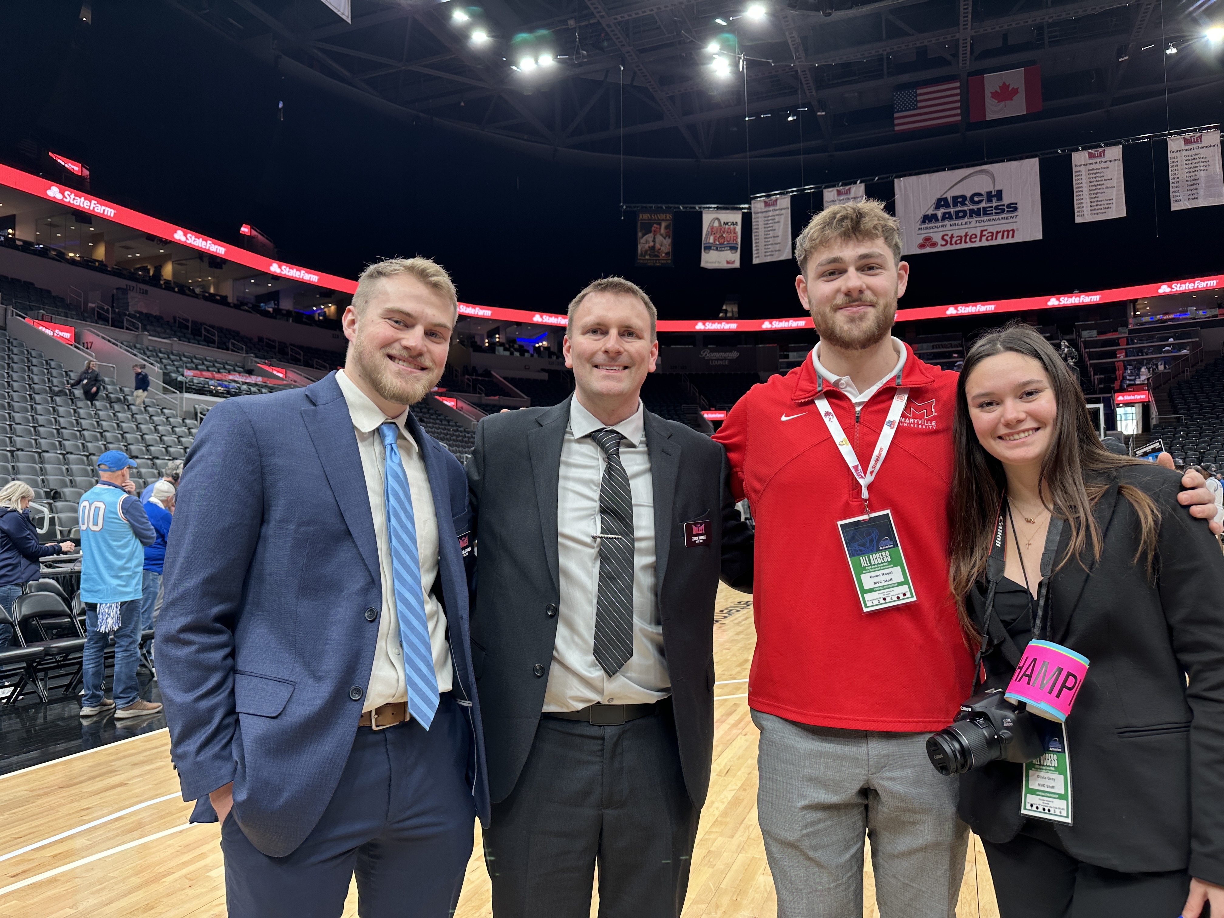 Three White men and a White woman stand courtside at a basketball arena, smiling for a photo. Two men wear suits, one wears a red shirt, and the woman holds a camera with an event badge. Behind them, banners for “Arch Madness” and “State Farm” hang above partially filled stands.