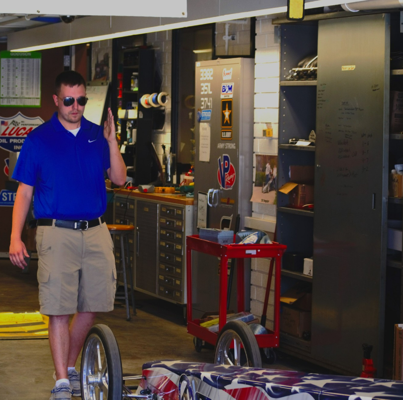 A white man wearing sunglasses, a blue collared shirt, and khaki shorts walks beside a small race car in a workshop filled with tools, shelves, and equipment.