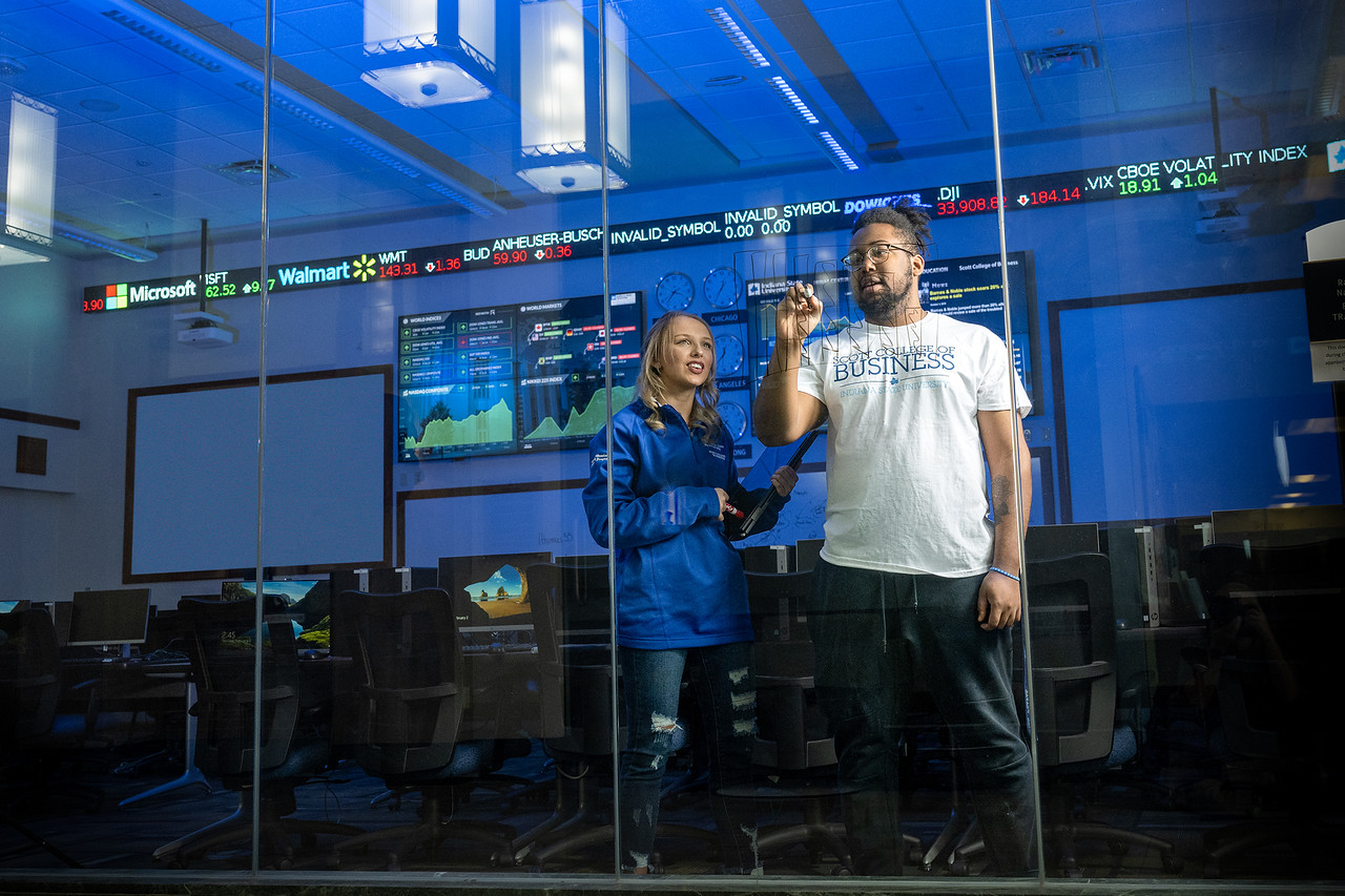 A Black man wearing glasses and a white “Scott College of Business” T-shirt stands beside a white woman in a blue jacket inside a computer lab. He writes on a glass wall while she watches. Digital tickers and large monitors displaying data are visible in the background.