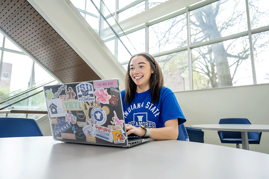 A cheerful female student works on her laptop at a table, engaged in her studies.