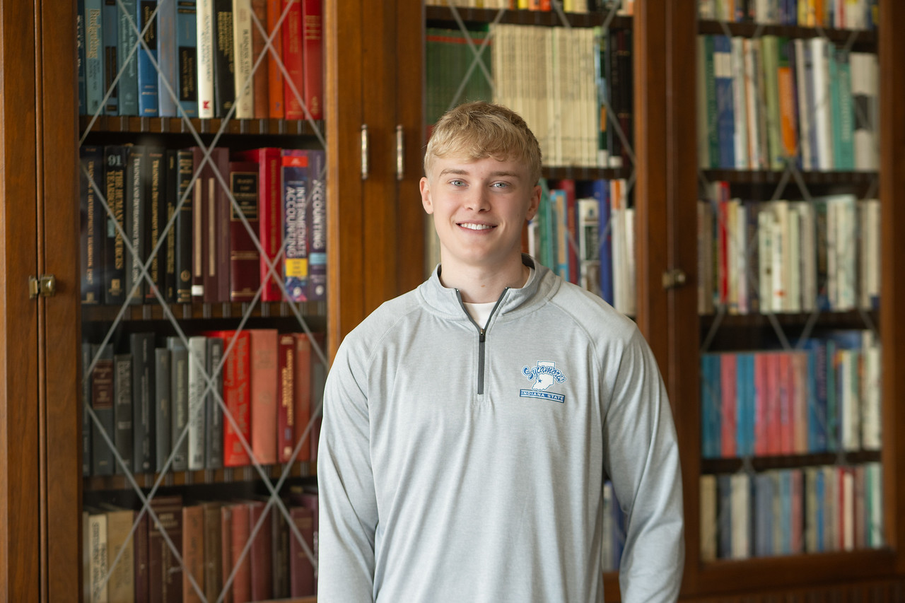 A white male with short blond hair wearing a light gray quarter-zip pullover stands in front of a large wooden bookshelf filled with colorful books. The shelves have glass doors with a crisscross design, and the setting appears to be a library or study room.