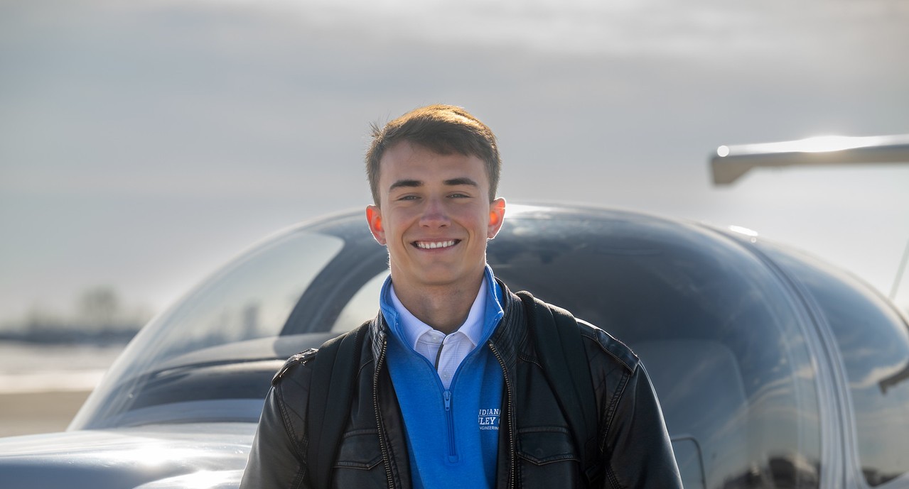 A white male wearing a blue quarter‑zip sweater, white collared shirt, and black leather jacket stands in front of a small aircraft on a bright, partly cloudy day.
