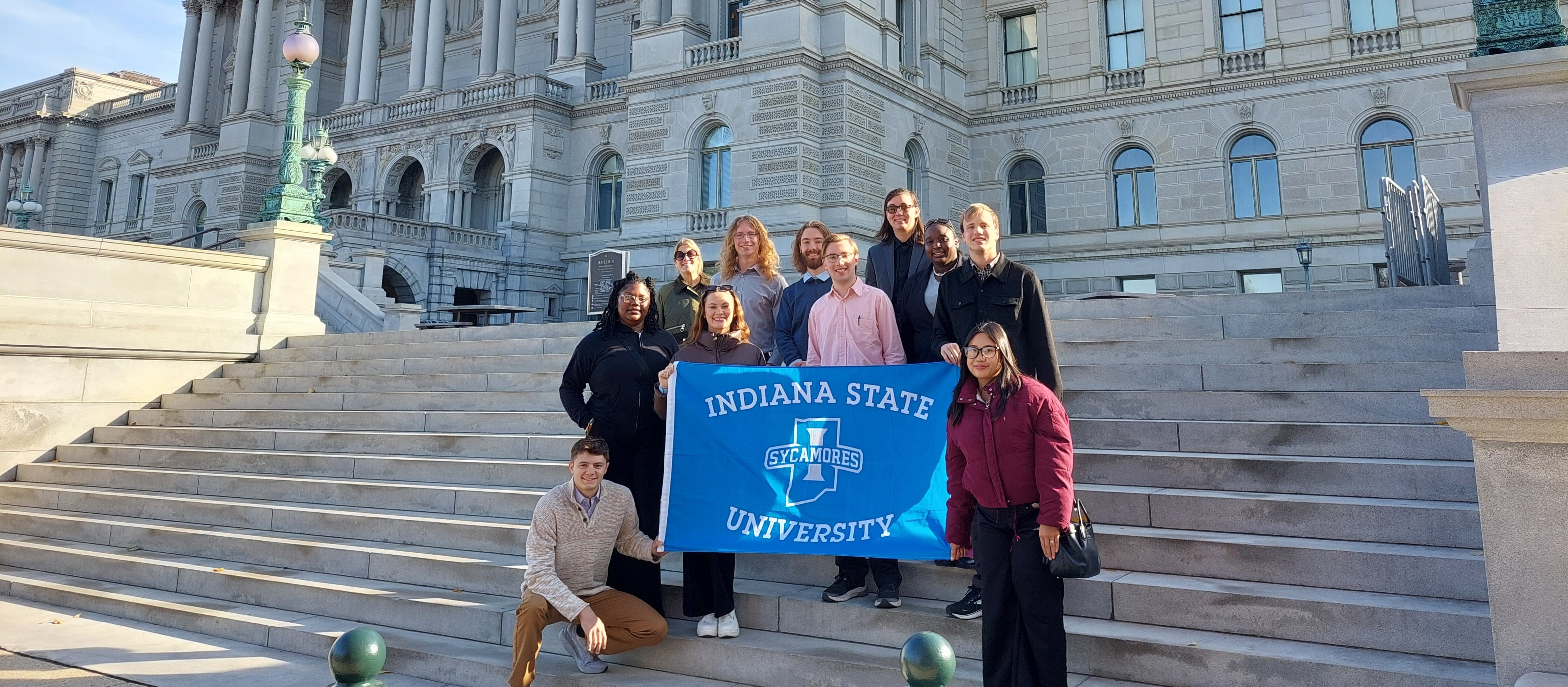 A group of people stands together on wide stone steps in front of a large historic building with ornate columns and arched windows. They hold a bright blue banner that reads “Indiana State University Sycamores.” The sky is clear and sunny, and the group poses smiling for the photo.