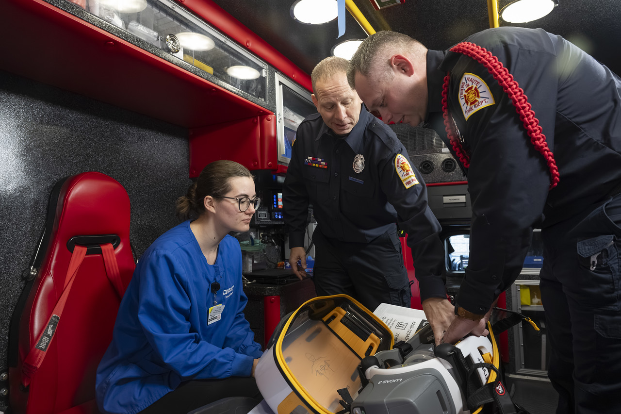 Inside an ambulance, two emergency responders stand beside a seated healthcare worker in blue scrubs, reviewing medical equipment and documentation in a brightly lit, red‑and‑black interior.