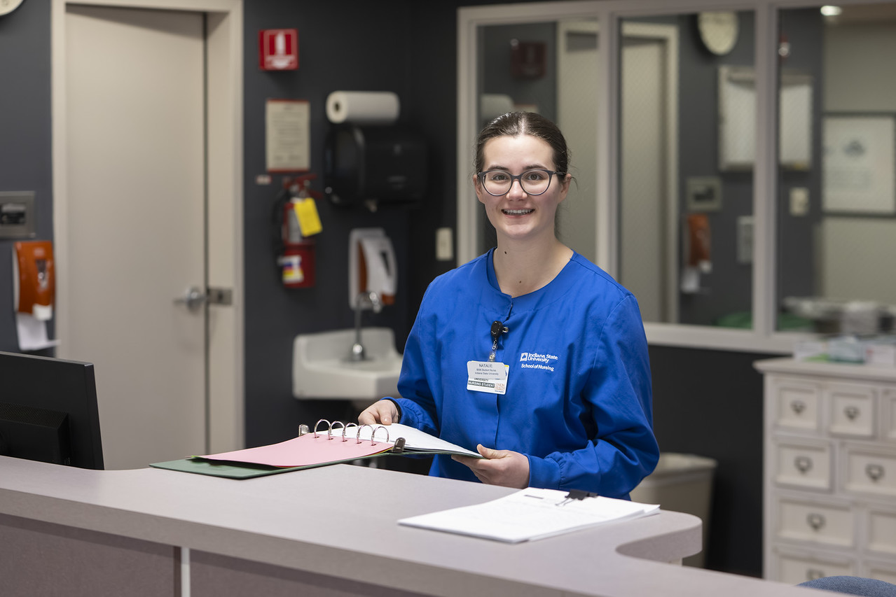 A white female student with brown hair pulled back into a ponytail wears blue nursing scrubs. She stands behind a reception desk in a healthcare setting holding an open binder with documents. Sinks, cabinets, and medical supplies are visible in the background.
