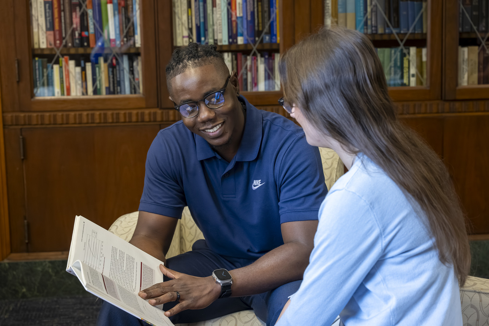 Student and tutor sitting together in a library, reviewing an open textbook and discussing the material in front of bookshelves.