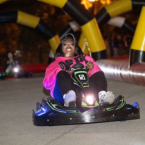 A person riding a low, electric go-kart–style vehicle on an indoor track. The rider is wearing a bright pink hoodie and sneakers, with legs extended forward toward the front of the kart. The vehicle has glowing blue and white lights along its base and a visible number “5” on the front panel. The track is bordered by padded barriers, and the scene appears to be lit for an entertainment or arcade setting.