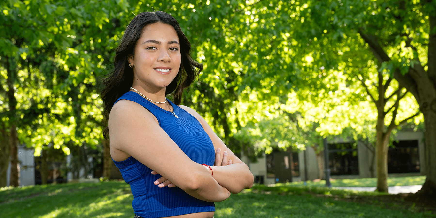 Smiling young woman with shoulder-length black hair standing outdoors in a grassy campus area with leafy trees. She wears a blue sleeveless top and is standing with arms crossed.