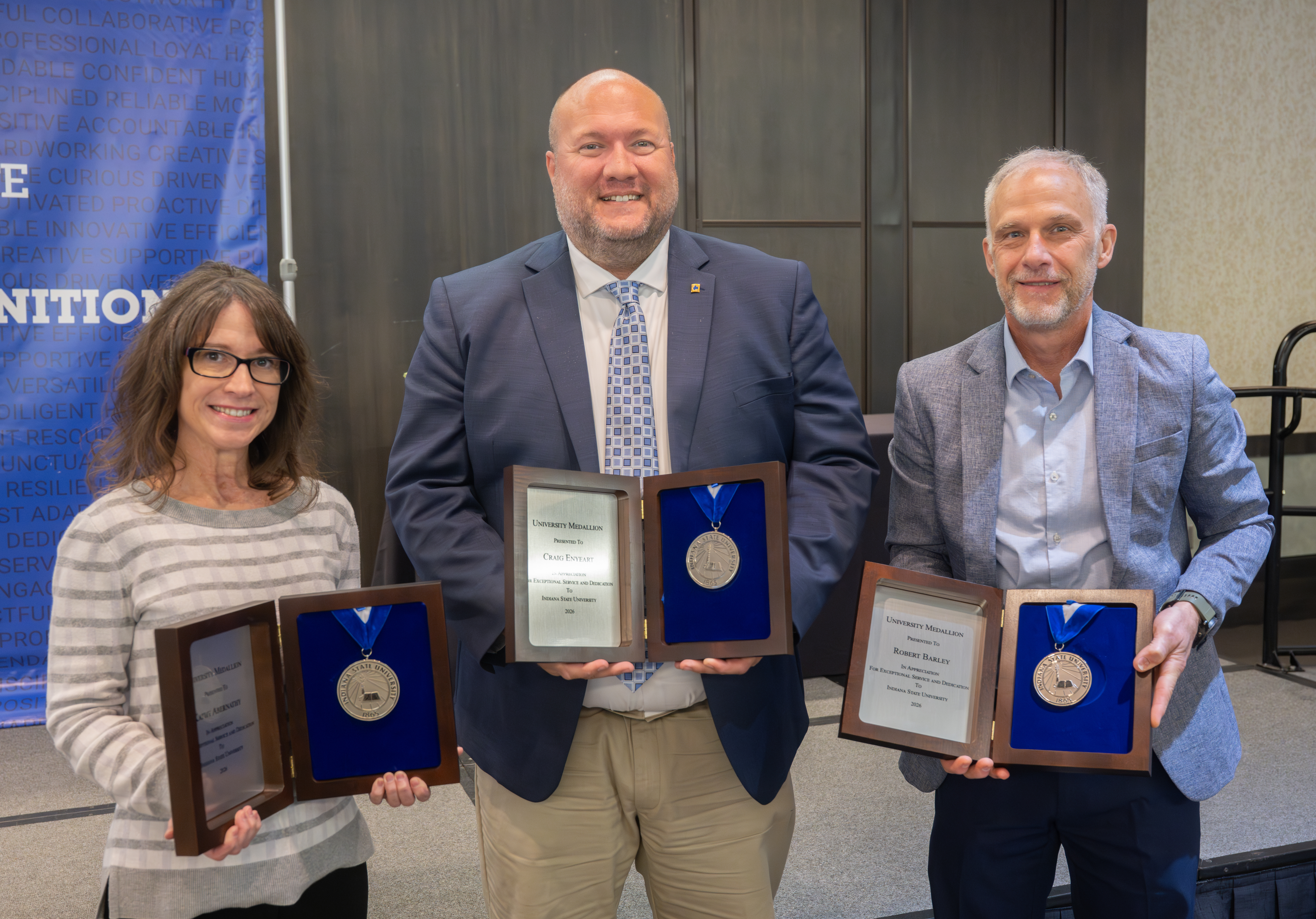 Three people holding university medallion awards and plaques while standing at an indoor recognition event.