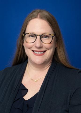 Professional headshot of a person with long straight hair wearing round glasses and a dark blazer over a blouse, smiling against a solid blue studio background.