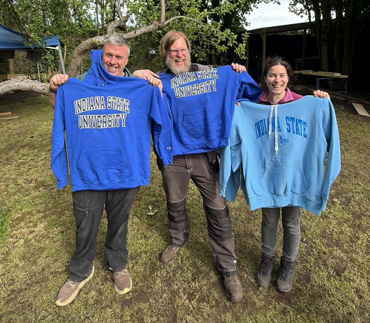 Three people standing outdoors, each holding up a blue Indiana State University hoodie. They are smiling in a grassy area with trees and a building in the background.