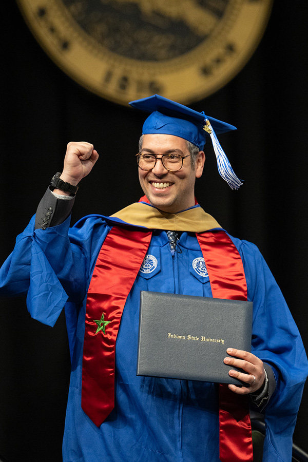 A smiling graduate in a blue cap and gown raises a fist in celebration while holding a diploma cover during a commencement ceremony. The graduate wears glasses, a gold hood, and a red stole, standing against a dark stage backdrop with a blurred university seal above.