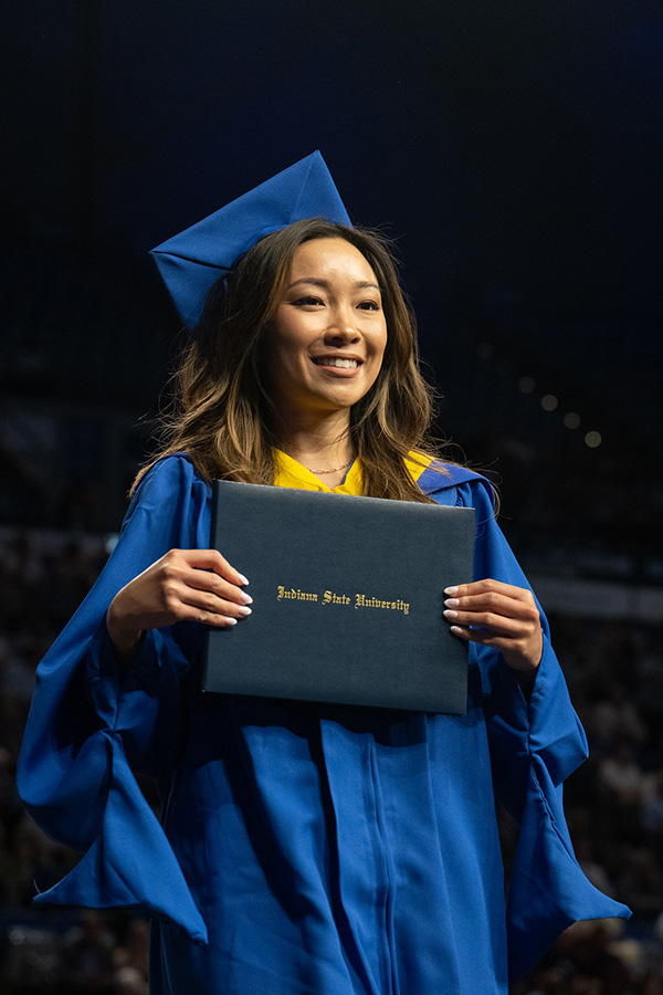 A graduate in a blue cap and gown smiles while holding a diploma cover during a commencement ceremony. The graduate stands onstage under dim lighting, with the audience blurred in the background.