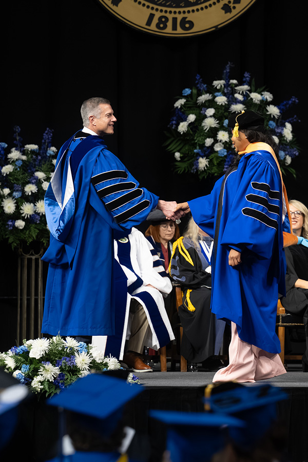 A university official in blue academic regalia shakes hands with a graduate wearing a blue doctoral gown and cap onstage during a commencement ceremony. Floral arrangements and seated faculty members are visible in the background, with graduates in blue caps in the foreground.