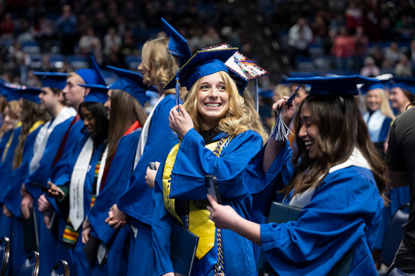A line of graduates in blue caps and gowns stand together at a graduation ceremony in a large indoor arena. In the center, a smiling graduate with decorated cap and honor cords adjusts her cap while holding a diploma cover. Another graduate in the foreground appears to be taking a photo or checking a phone. The background shows a crowd of attendees seated in dim lighting.