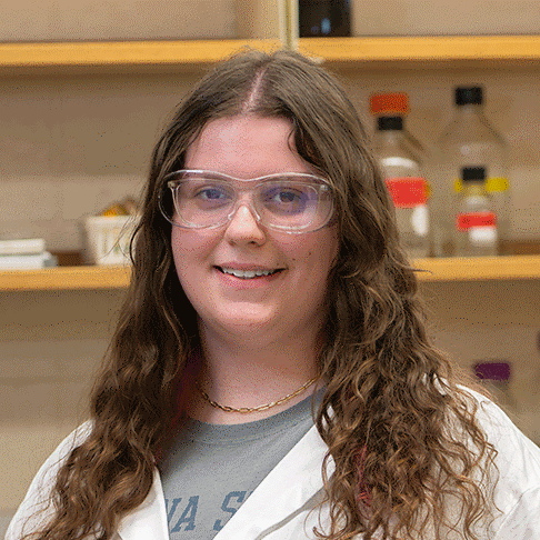 A white female student with curly brown hair wearing safety goggles and a lab coat standing in a science lab, with shelves of bottles and lab equipment in the background.