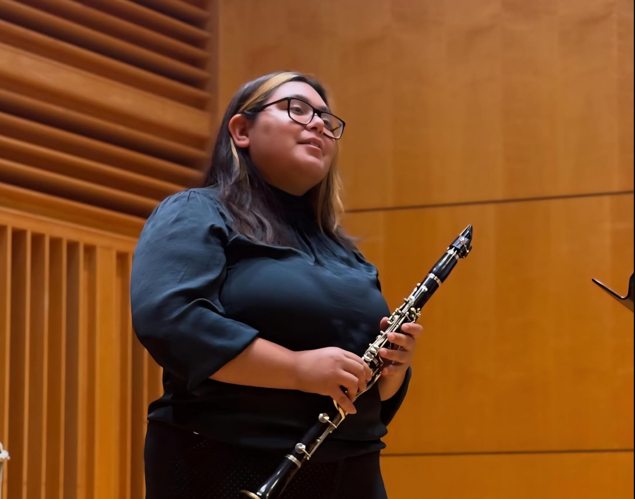 A woman with dark brown hair stands in a wood-paneled performance hall holding a clarinet, facing slightly to the side. She wears a dark black dress shirt and matching pants. 