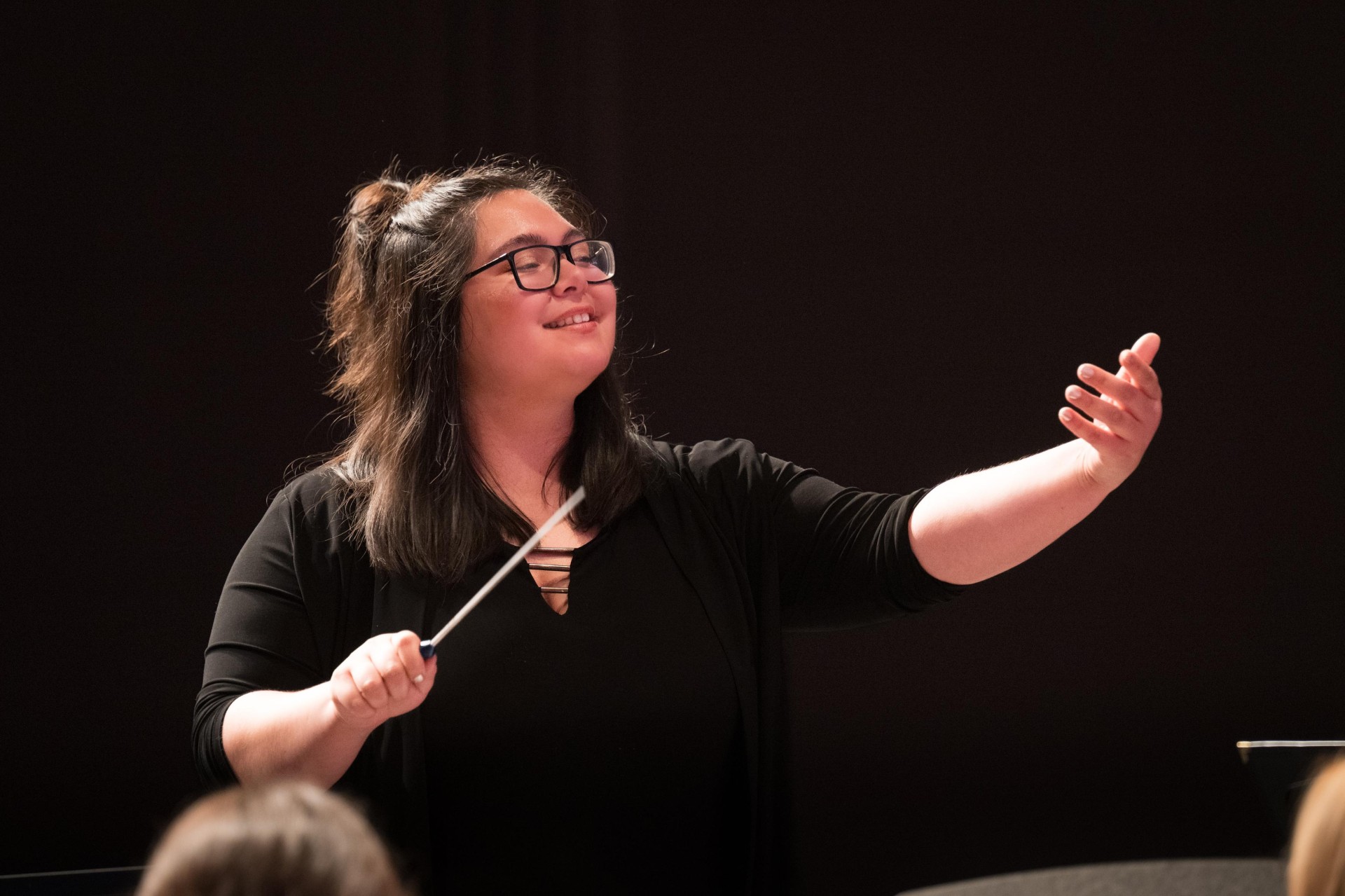 A woman with dark brown hair conducts a music performance, holding a baton and raising one hand, standing in front of a dark stage background. She wears a black dress shirt. 