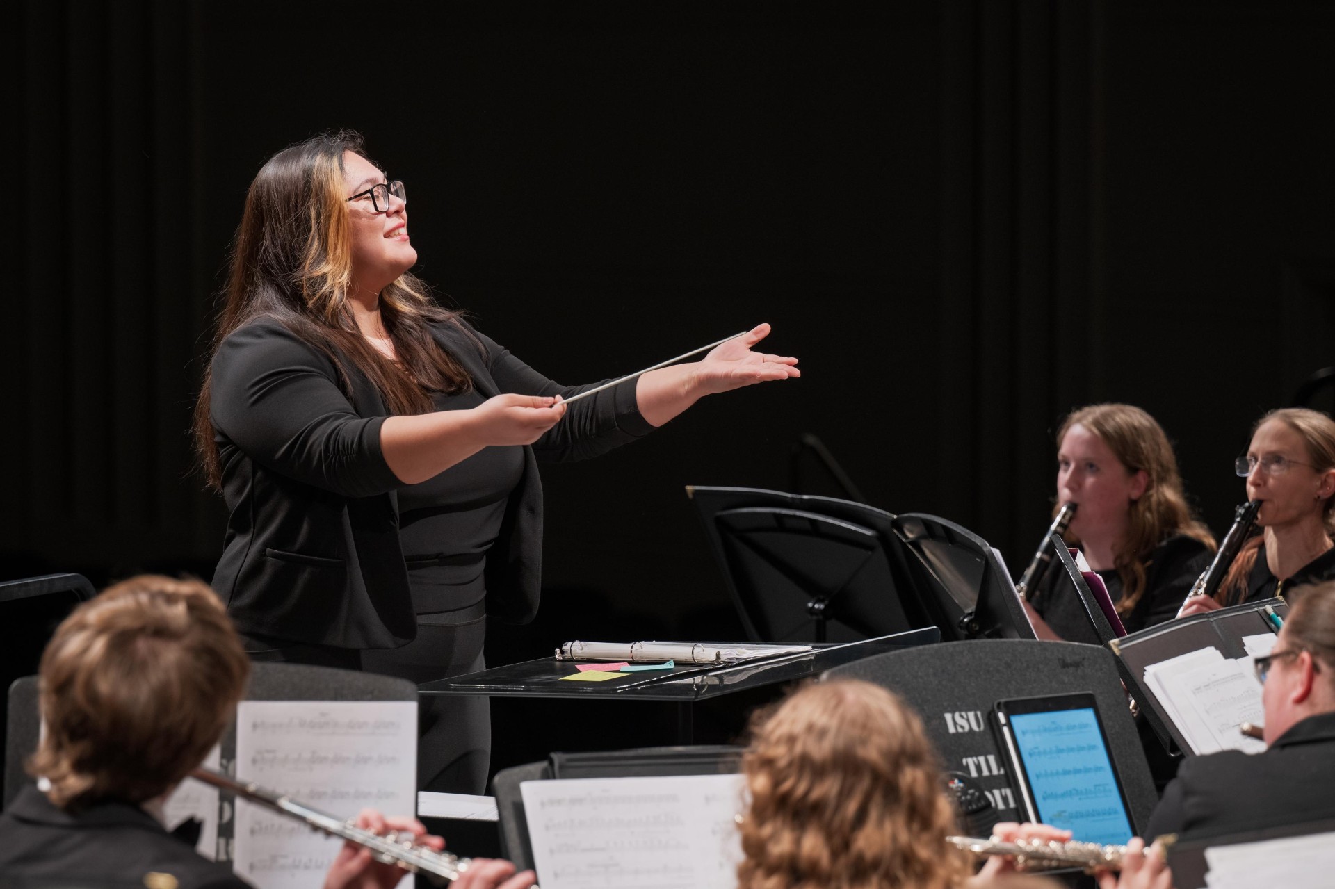 A woman with dark brown hair conducting a music performance, holding a baton and raising one hand toward an ensemble of musicians. 
