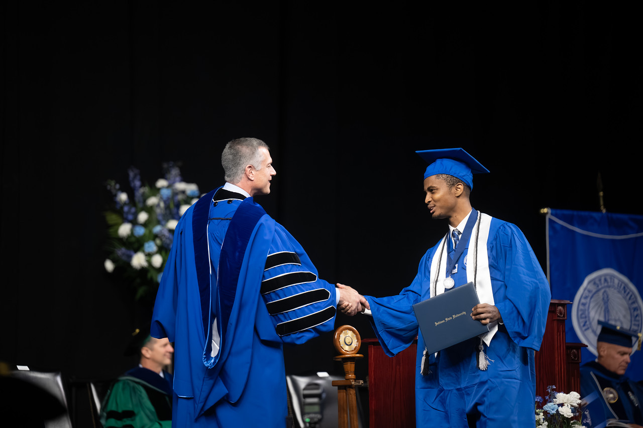 Young Black male in a blue cap and gown shakes hands with a Indiana State University President Dr. Mike Godard on stage while holding a diploma. The graduate wears a white stole, honor cords, and a medal. A podium, seated faculty, and stage decorations are visible in the background.