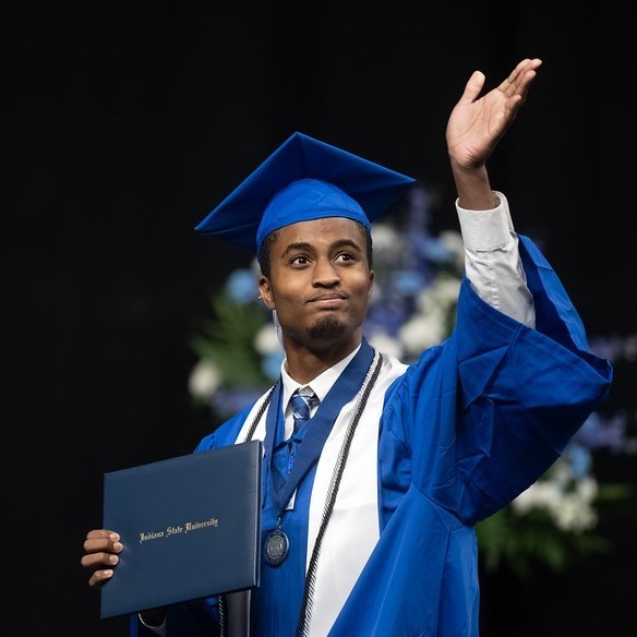 Young Black male in a blue cap and gown holds a diploma and raises one hand while standing on stage at a commencement ceremony. The graduate wears a white stole, honor cords, and a medal. An American flag and seated faculty are visible in the background.