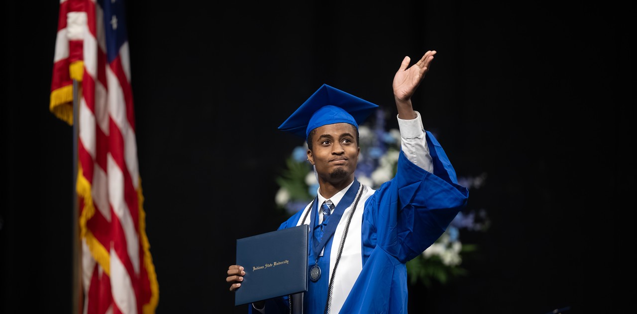 Young Black male in a blue cap and gown holds a diploma and raises one hand while standing on stage at a commencement ceremony. The graduate wears a white stole, honor cords, and a medal. An American flag and seated faculty are visible in the background.