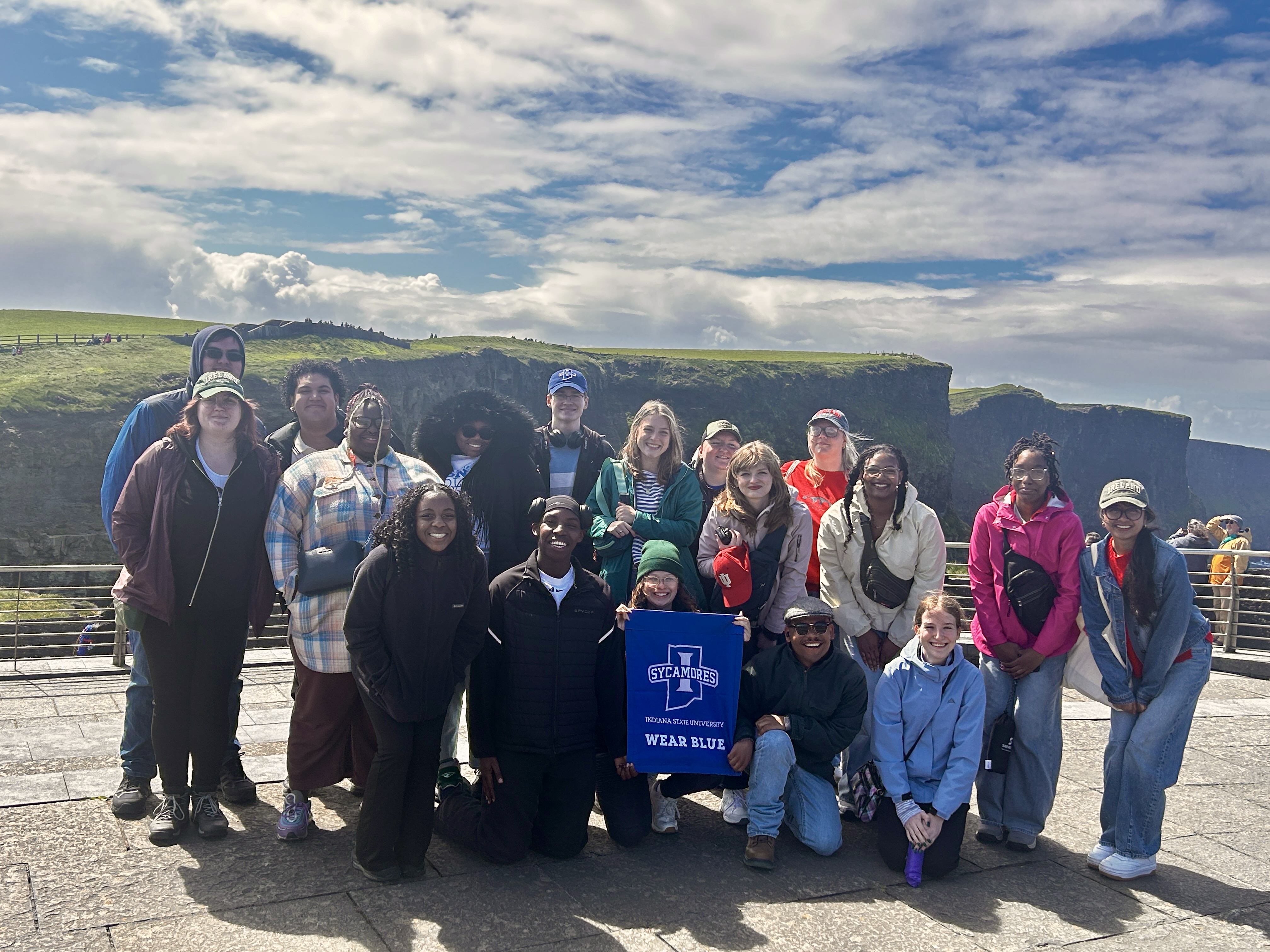 A group of Indiana State Students smiling in front of the Cliffs of Moher in Ireland. One individual is holding a Indiana State University sign. The sky is partly cloudy, and the landscape features green cliffs and ocean views in the background.