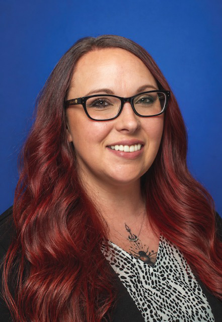 The image shows a person in a professional studio-style headshot against a blue background. They have long wavy dark red hair, black rectangular glasses, and are smiling. They’re wearing a black blazer over a patterned blouse, along with a visible chest tattoo and necklace. The overall presentation looks polished and suitable for a business profile, company directory, or LinkedIn-style portrait.
