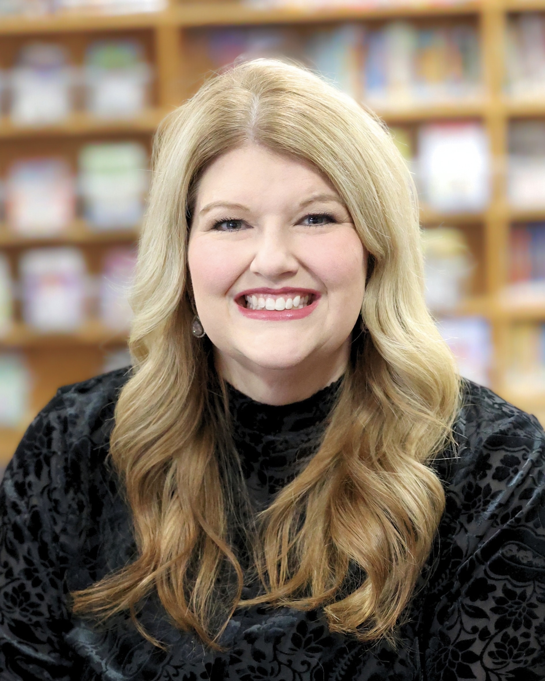 The image is a warm, professional portrait of a smiling person seated indoors. They have long blonde hair styled in loose waves and are wearing a black patterned top with earrings. The background is softly blurred and appears to be bookshelves or a library/bookstore setting, giving the portrait an approachable academic or professional atmosphere.