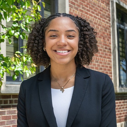 A Black female student wearing a dark blazer and white shirt stands outdoors near a brick building and greenery, facing the camera.
