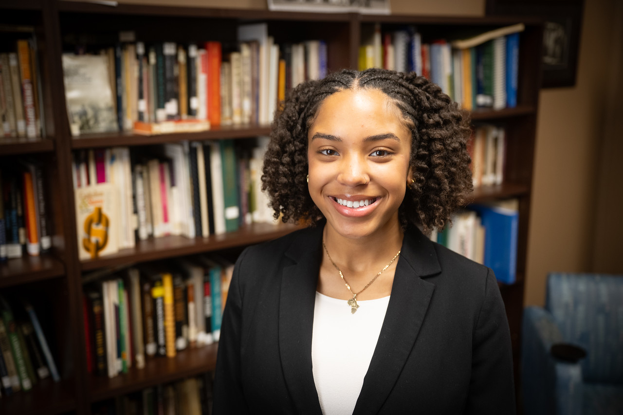Black female student in a black blazer and light-colored under shirt standing in front of a bookshelf filled with books in an indoor study or office setting.
