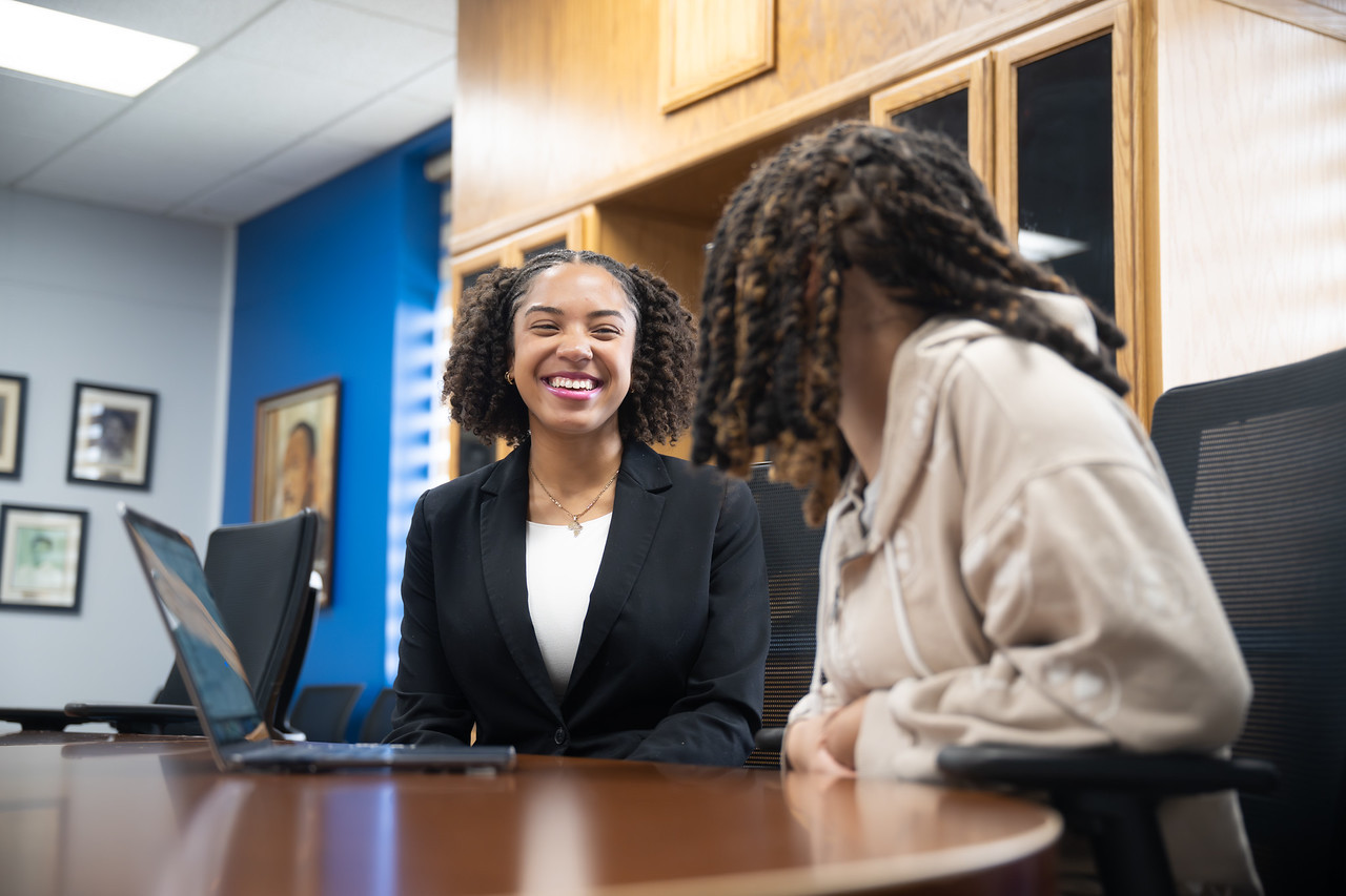 Black female student in a black blazer and a light-colored under shirt sitting at a table with a laptop, speaking with another Black student in a campus office or meeting room.