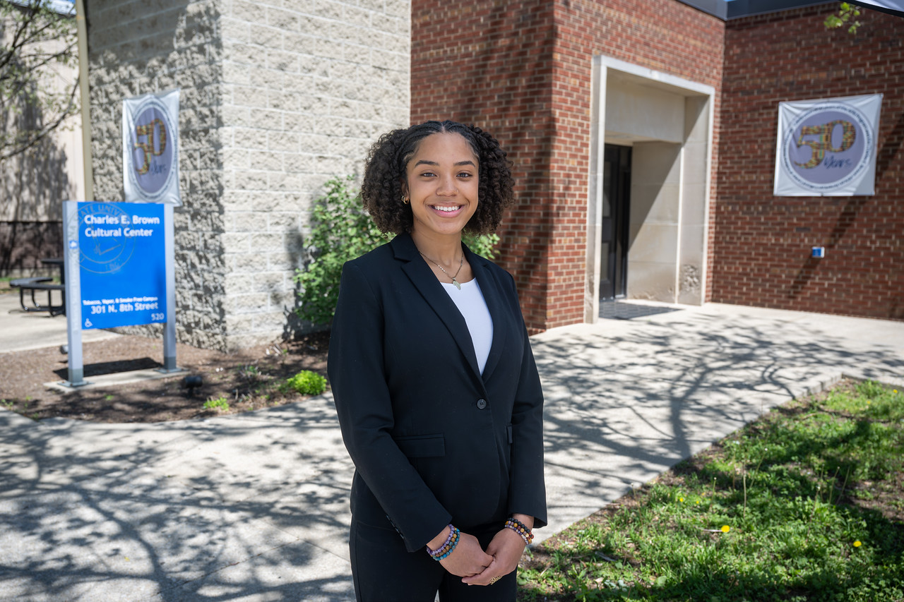 Black female student in a black blazer and a light-colored under shirt standing outside a brick campus building near a sign for the Charles E. Brown Cultural Center.