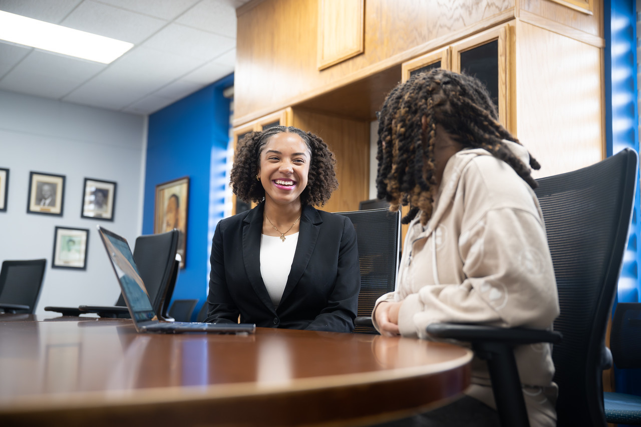 Two people are seated at a conference table in an office or meeting room. One person in a black blazer is smiling and appears engaged in conversation with the other person, who is turned slightly away from the camera. A laptop is open on the table, and the room has wood cabinetry, framed photos on the walls, and blue accent walls. The atmosphere appears professional and friendly.