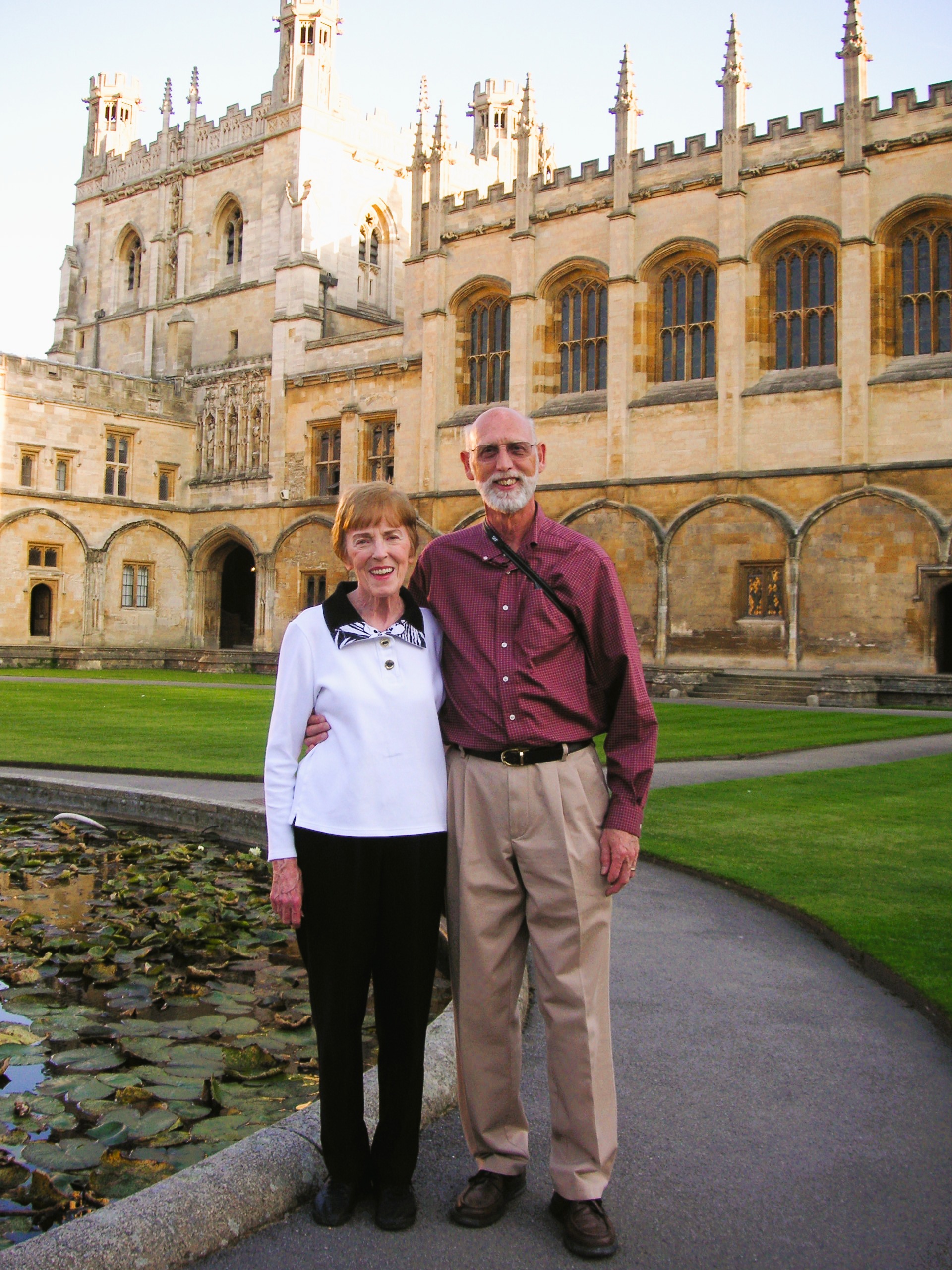 Two people standing together in front of a historic stone university building with gothic architecture and a courtyard pond.
