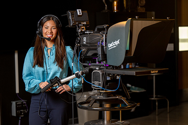 A smiling young woman with dark hair and wearing a headset stands behind a professional broadcast camera mounted on a tripod inside a television production studio.